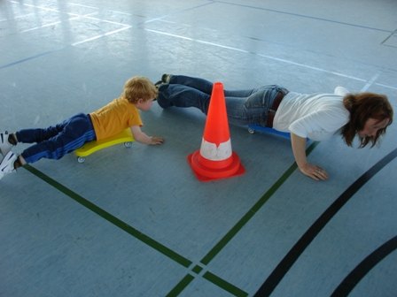 Kind und Erwachsene bewegen sich mit Rollbrett und Hütchen in der Sporthalle